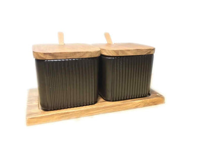 Two black ceramic canisters with wooden lids on a wooden tray against a white background