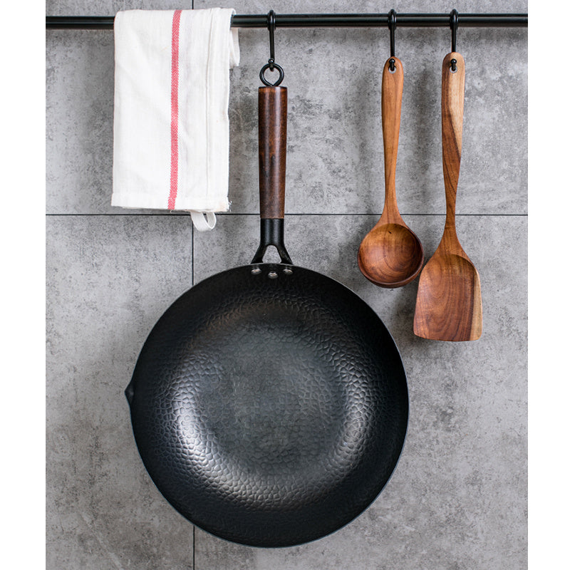 Black wok with wooden handle and two wooden spoons hanging on a metal rod against a gray tiled wall.