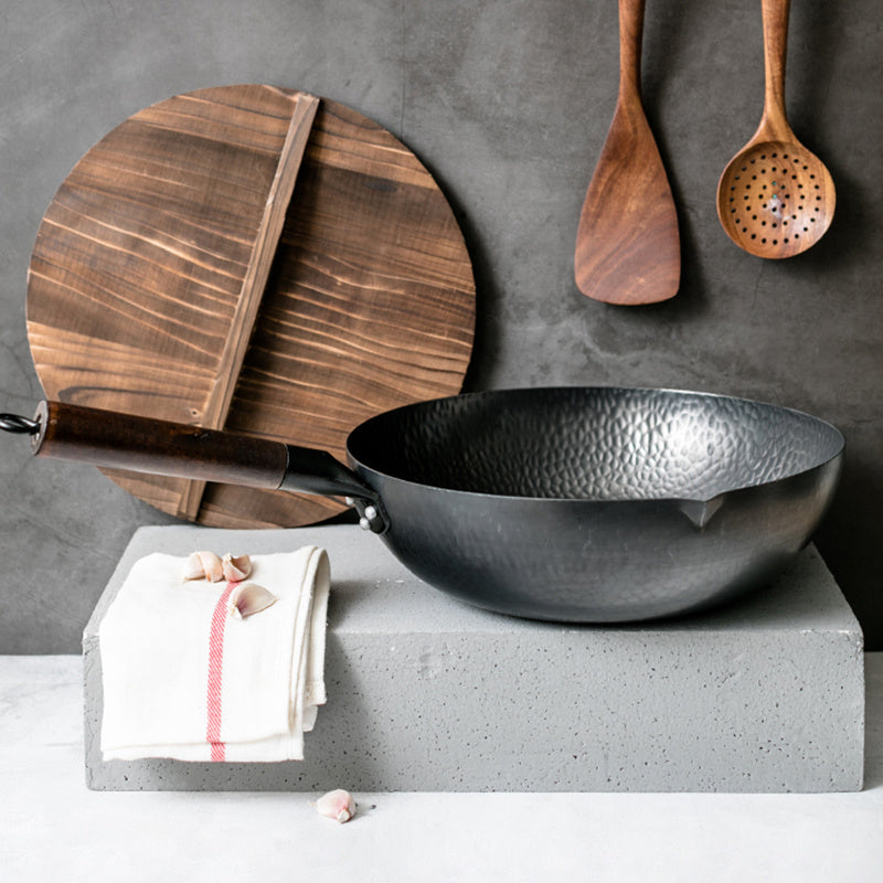 Black wok on a concrete block with wooden cutting board, spoons, and towel against a gray background