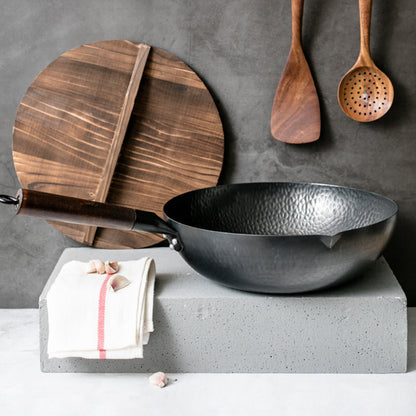 Black wok on a concrete block with wooden cutting board, spoons, and towel against a gray background