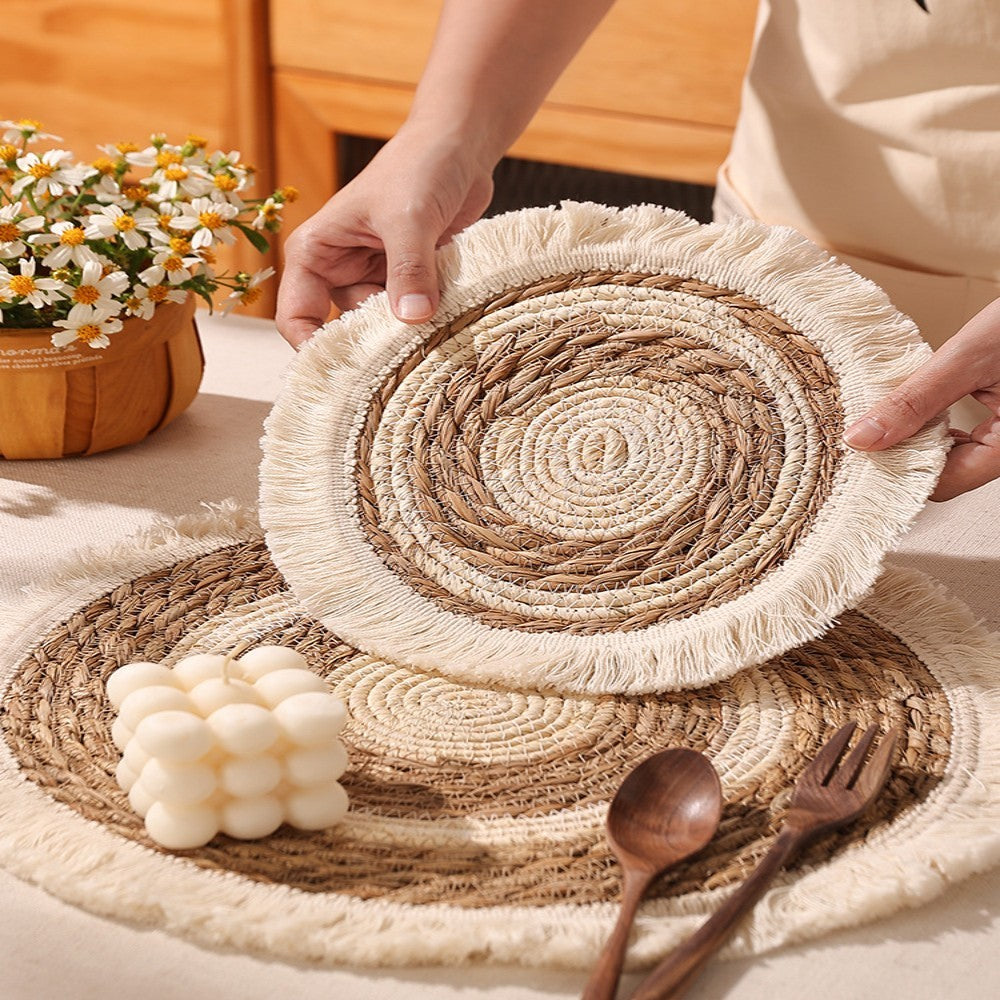 Woven placemats with fringe held by a person, wooden cutlery and flowers in the background.