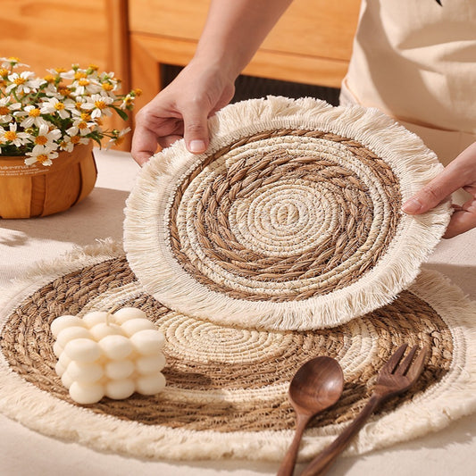 Woven placemats with fringe held by a person, wooden cutlery and flowers in the background.