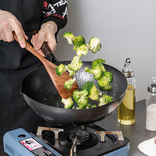 Person cooking broccoli in a wok on a portable stove with a wooden spatula.